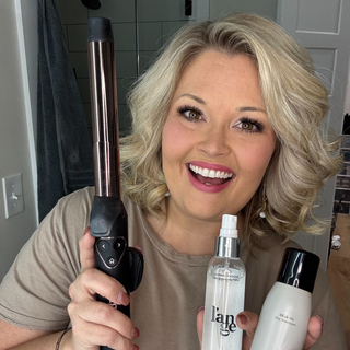 Smiling woman holding a curling iron, thermal protection spray, and dry shampoo, showcasing hairstyling and care products.