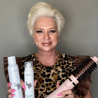 Woman with short blond hair holding dry texture spray, hair boost primer, and a pink brush, wearing a leopard print top and gold chain necklace.