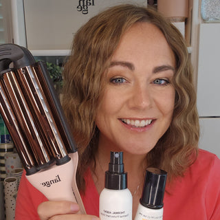 A woman smiling while holding a hair crimper and two hair care products, showcasing styling tools and products.