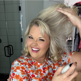 Smiling person with blonde curls styles hair using a spray, wearing a floral blouse in a bright interior setting.