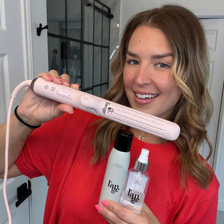 Person showcasing a pink hair styling tool alongside two hair care products, including a dry shampoo and a spray, against a bathroom backdrop.
