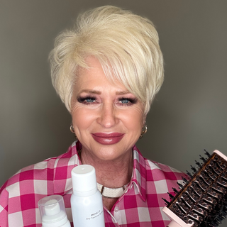 Smiling person with short platinum blonde hair holds styling products and a round brush, wearing a pink checkered shirt.