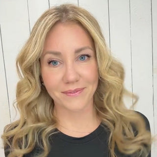 Woman with long, wavy blonde hair against a white wooden backdrop, wearing a black top and natural makeup.