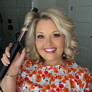 Smiling woman with curled, blonde hair using a curling iron, wearing a floral-patterned blouse.