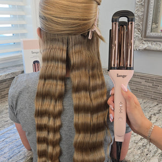 Styling tool creating defined waves in long, blonde hair, held by a hand with manicured nails against a salon backdrop.