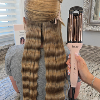 Styling tool creating defined waves in long, blonde hair, held by a hand with manicured nails against a salon backdrop.