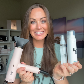 Smiling woman showcasing a hair dryer and two hair care products in a well-lit room. Shelves with additional products are visible in the background.