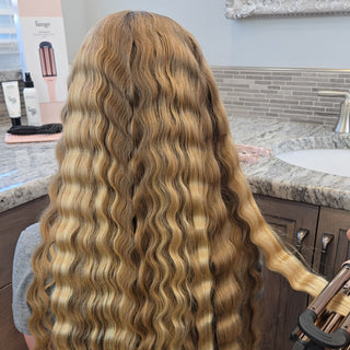 Long, wavy blonde hair styled with a curling iron near a bathroom counter featuring styling products and tools.
