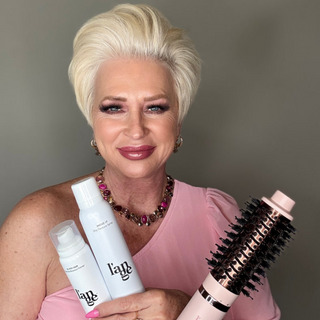 Woman with short blonde hair holding two hair care products and a styling tool, wearing a pink top and matching jewelry, smiles at the camera.