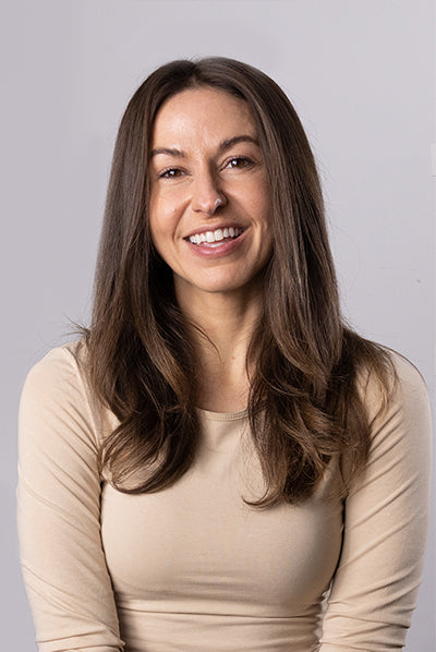 A smiling woman with long brown hair wearing a beige top, set against a plain background.