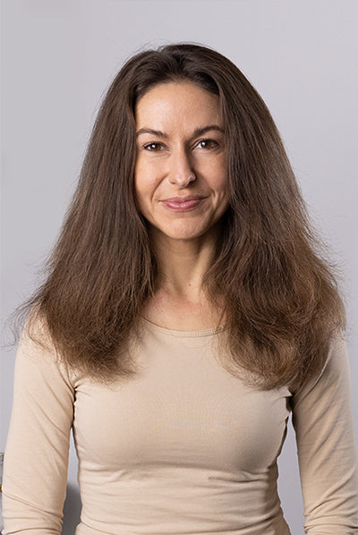 A person with long, voluminous brown hair wearing a beige top smiles against a plain background.
