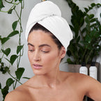 A dark-haired woman in a bathroom wears the white microfiber hair towel on her hair.