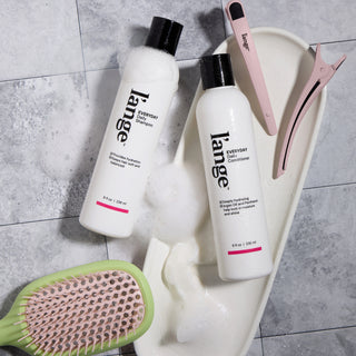 Overhead view of L’ange Shampoo and Conditioner with a hairbrush and clips on a grey tile surface