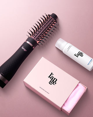 A black and rose-gold hair styling tool, a white bottle of heat shield spray, and a pink box containing a microfiber hair wrap are arranged against a soft pink background.