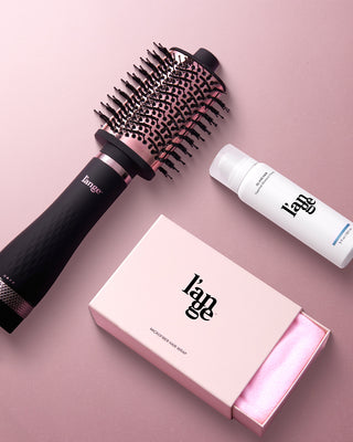 A round hair brush, a bottle of styling mousse, and a pink microfiber hair wrap in branded packaging are arranged on a soft pink surface.