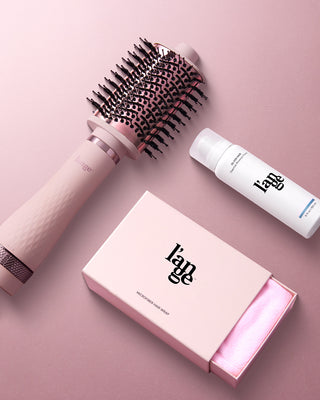 Pink hair styling products on a pink background, including a round brush, a microfiber hair wrap in a box, and a styling mousse.