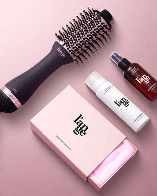 A collection of hair care products including a round brush, hair spray, thermal styling spray, and a microfiber hair wrap in a pink box are arranged on a soft pink background.