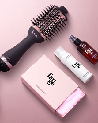 A hair styling set featuring a round brush, two hair care product bottles, and a pink microfiber hair wrap in a pink box against a soft pink background.