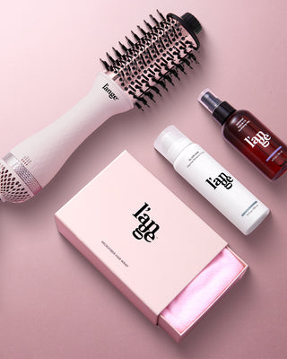 A collection of hair styling products including a round brush, spray bottle, mousse, and a boxed microfiber hair wrap, all arranged on a soft pink background.