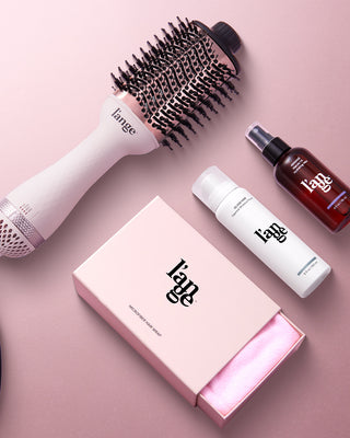 A collection of hair styling products includes a blow-dryer brush, a hair mist, a thermal protectant spray, and a microfiber hair wrap in a pink box, arranged on a soft pink surface.