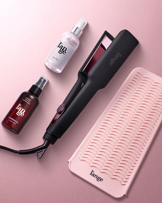 A collection of hairstyling products including a black flat iron, a bottle of hair spray, and a pink textured mat on a soft pink background.