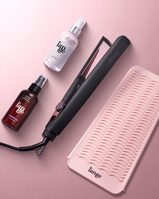 A sleek black hair straightener is accompanied by styling products and a textured pink heat-resistant mat on a soft pink background.