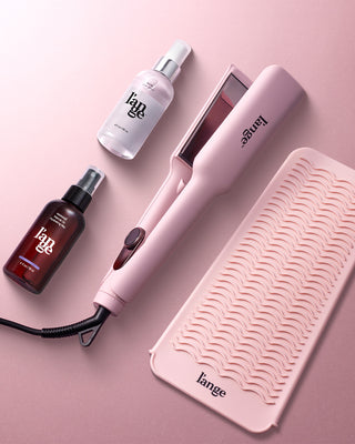 Pink styling tool set with two bottles of hair spray and a textured silicone mat on a matching pink background.