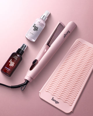 A collection of hair styling tools and products, including a pink flat iron, two spray bottles, and a wavy-textured heat mat, placed on a coordinating pink background.