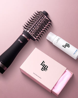 A styling brush, a bottle of thermal styling glaze, and a box containing a microfiber hair wrap are arranged on a soft pink surface.