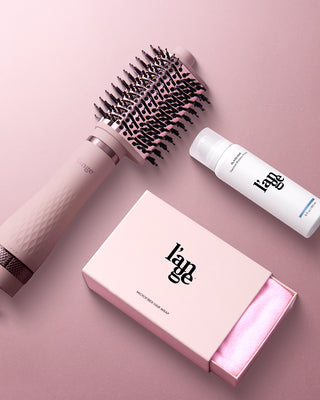 A stylish hair care set including a pink round brush, a white bottle of heat shield spray, and a boxed microfiber hair wrap, all displayed on a pink surface.