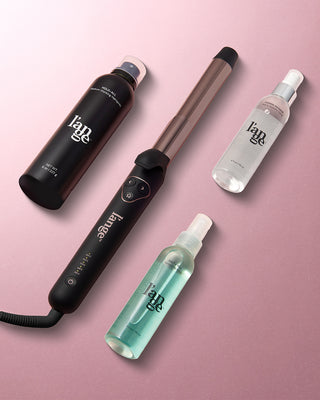 L'ange hair styling set featuring a black curling wand, volumizing mousse, heat shield spray, and sea salt texturizing mist, displayed on a pink background.