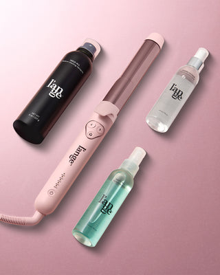 Pink curling wand alongside three hair styling products, including a black hold spray, a clear thermal spray, and a blue sea salt spray, arranged on a pink background.