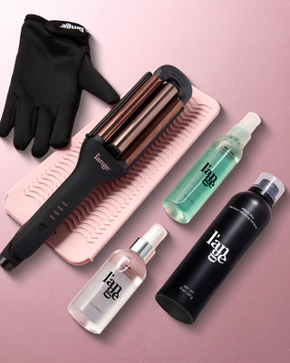Styling set featuring a black heat-resistant glove, a triple barrel curling iron on a pink mat, and three hair care products in sleek bottles arranged on a soft pink background.