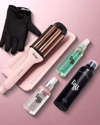 Hair styling set featuring a three-barrel curling iron on a pink mat, a heat-resistant glove, and a collection of styling sprays and products, displayed on a pink background.