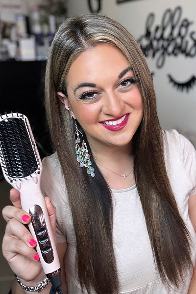 A woman with long, straight hair smiles while holding a hair styling brush, with a decorative background featuring a "hello gorgeous" sign.