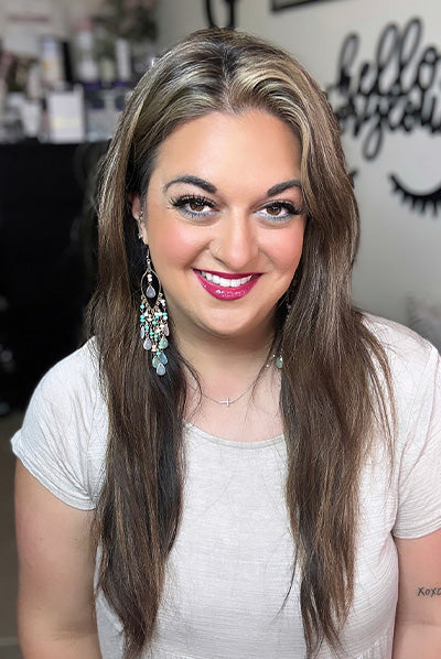 Smiling person with long brown hair wearing a white shirt and ornate earrings, with a blurred background featuring wall decor.