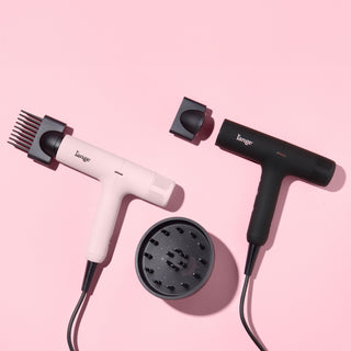 Two L’ange HauteAir hair dryers—one blush pink and one black—side by side with attachments  on a pink background 