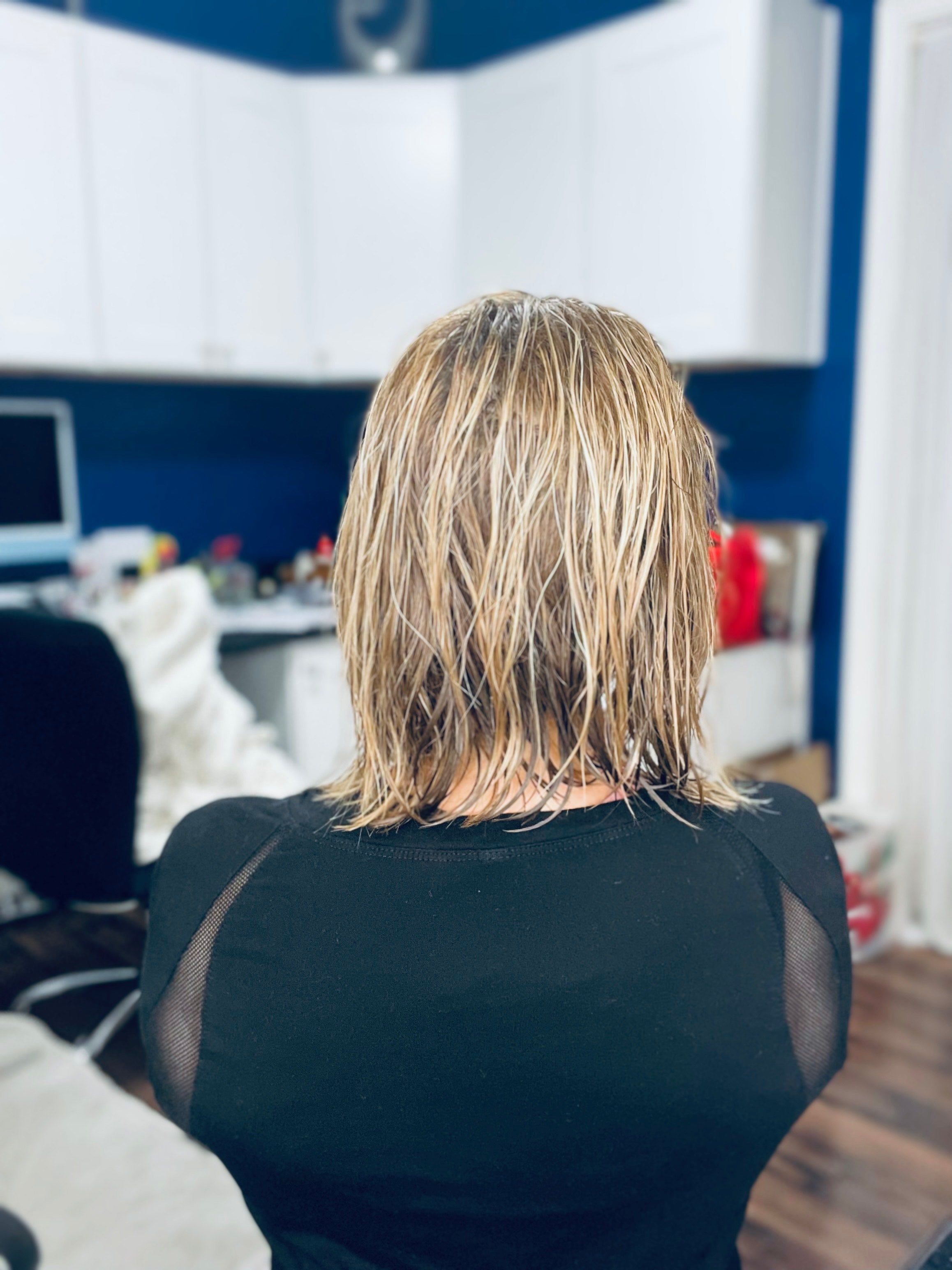 A person with wet, shoulder-length blonde hair is sitting in a modern office space with white cabinets and a computer in the background.
