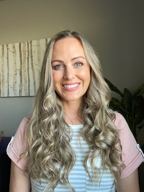 A person with long, wavy blonde hair and a striped shirt smiles warmly, standing in a room with a plant and a painting in the background.