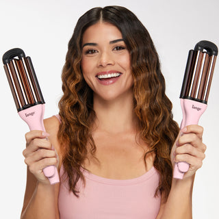 A smiling woman holds two pink, multi-barrel curling wands, showcasing her wavy hair.