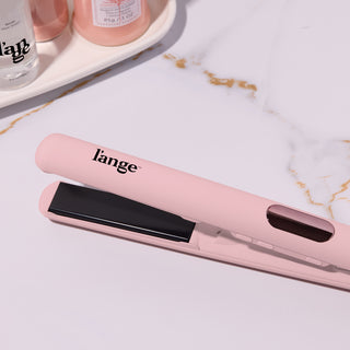 Pink flat iron with black plates, featuring the brand name on the handle, placed on a marble surface next to hair care products.