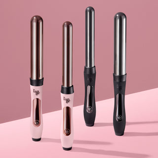 "Four L’ange curling wands standing upright on a pink surface with a pink background. Two are light pink with rose gold barrels and handles, and two are matte black with silver barrels and chrome accents.