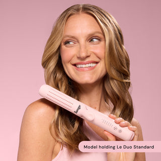 Smiling individual with wavy blonde hair showcasing the Le Duo Standard styling tool against a soft pink background.