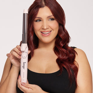 Smiling woman with long auburn hair holds a pink curling iron, showcasing its features.