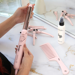 Pink styling essentials including a curling wand, spray bottle, comb, and hair clips arranged on a marble surface, showcasing beauty tools and products with a modern aesthetic.
