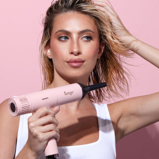 Woman using a pink l’ange hair dryer on damp, tousled hair against a pink background.