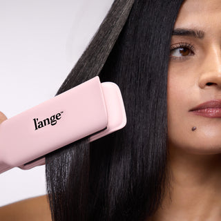 Close-up of a woman straightening her hair with a blush pink L’ange Le Titane flat iron. The tool glides through her shiny, straightened hair, highlighting product performance