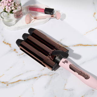 Blush triple-barrel hair waver on a marble countertop next to beauty accessories.
