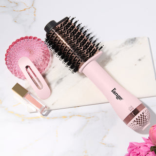 A pink L’ange hair dryer brush with black bristles placed on a marble surface, styled with a pink hair clip, pink glass dish, lip gloss, and pink flowers.