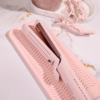 Pink hair straightener placed on a textured silicone mat, accompanied by two matching hair clips, set on a marble surface.
